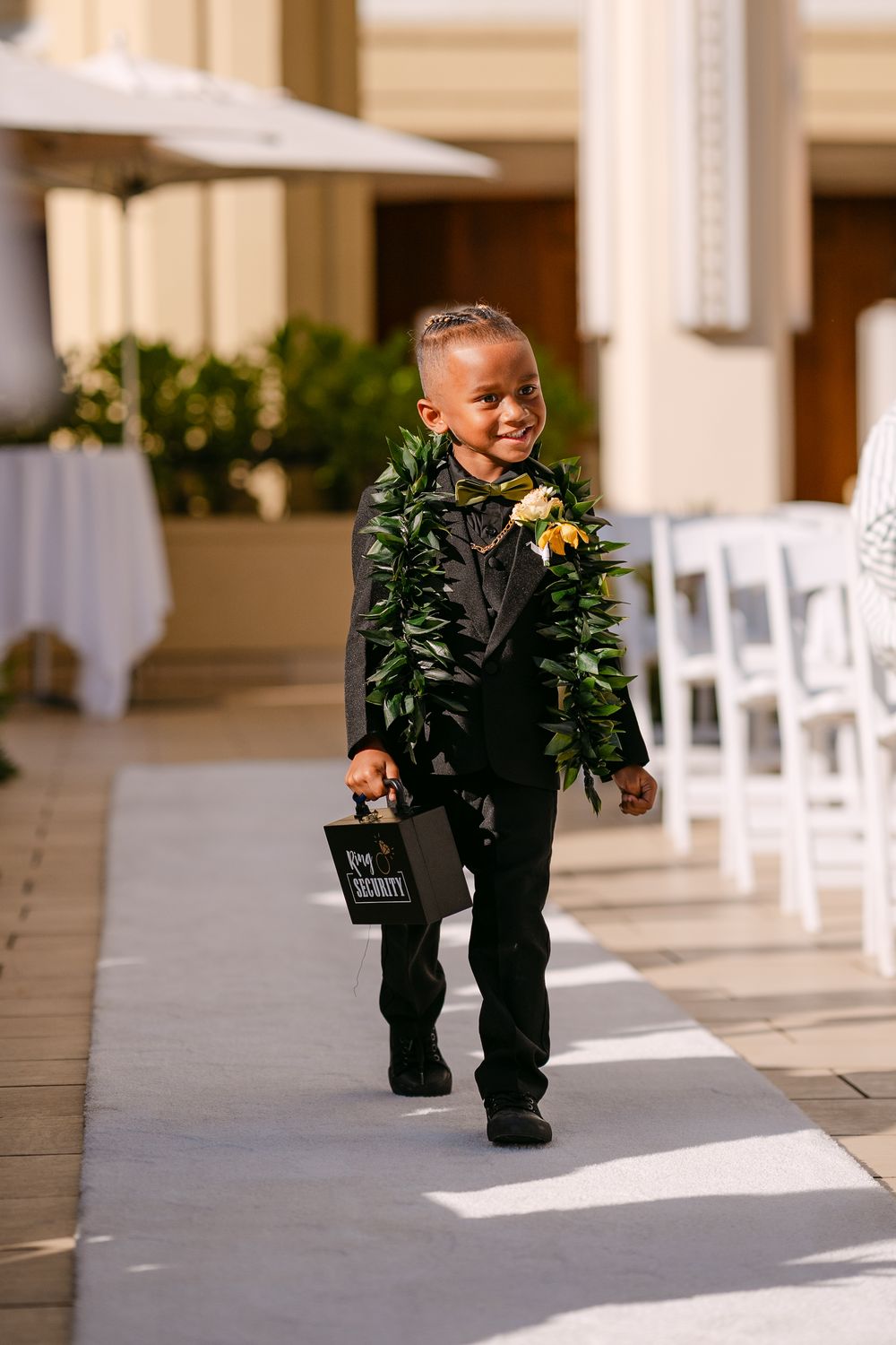 Young ring bearer in black tuxedo with floral lei walks down wedding aisle carrying small bag.