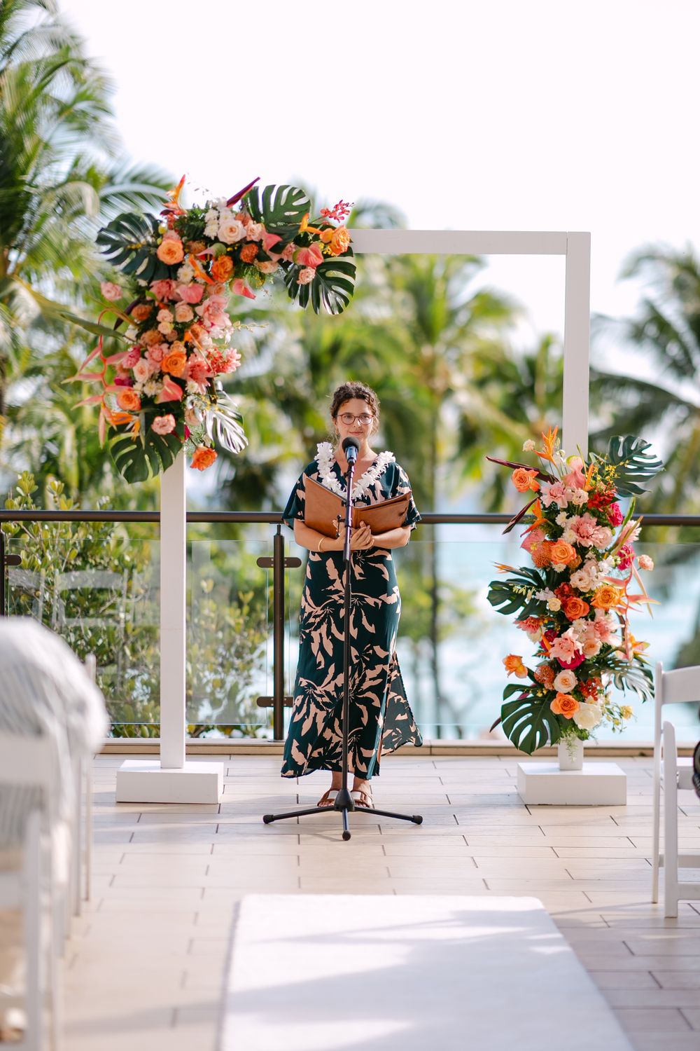 Officiant in floral dress stands at tropical outdoor wedding altar with orange blooms and monstera leaves.