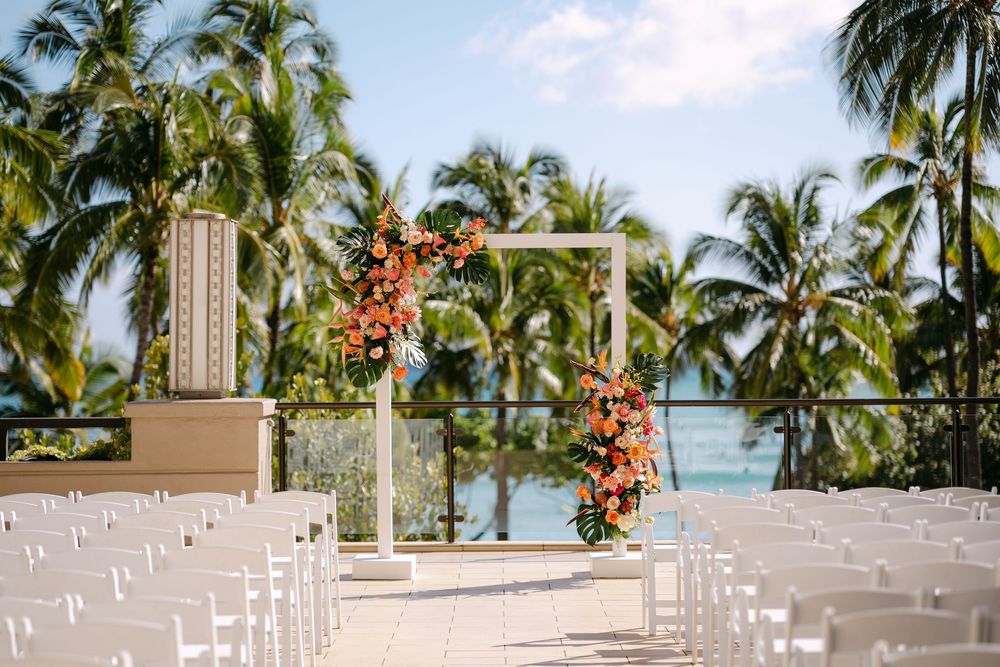 Tropical outdoor wedding ceremony setup with white chairs, floral arch, and palm trees overlooking the ocean.