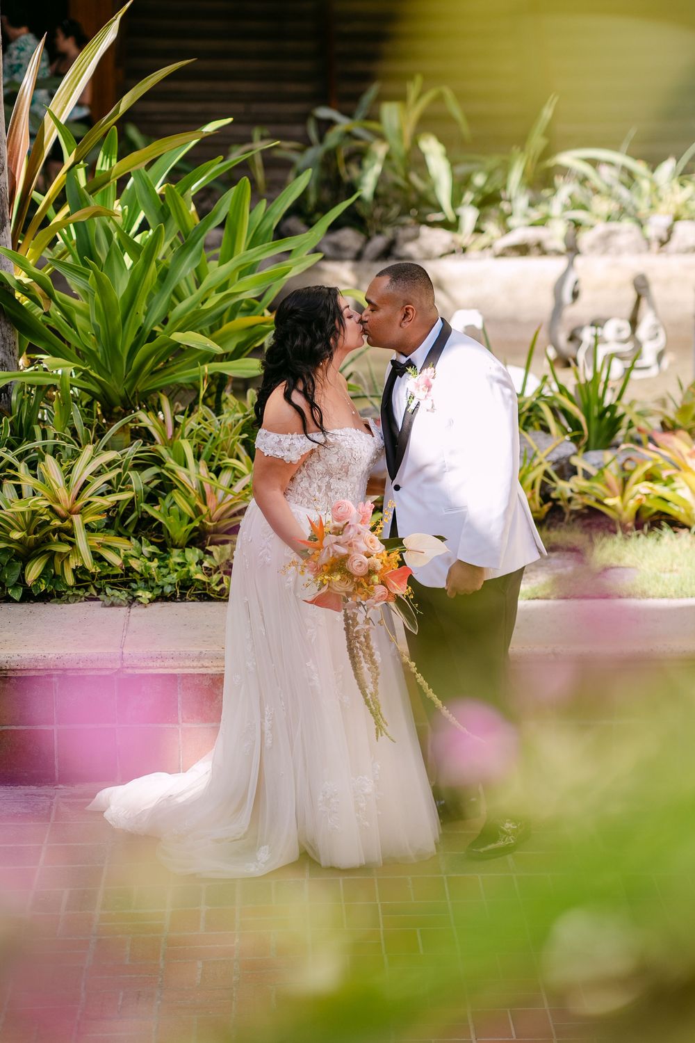 Bride and groom share a romantic kiss in a lush tropical garden, bride holding a colorful bouquet in a white gown.