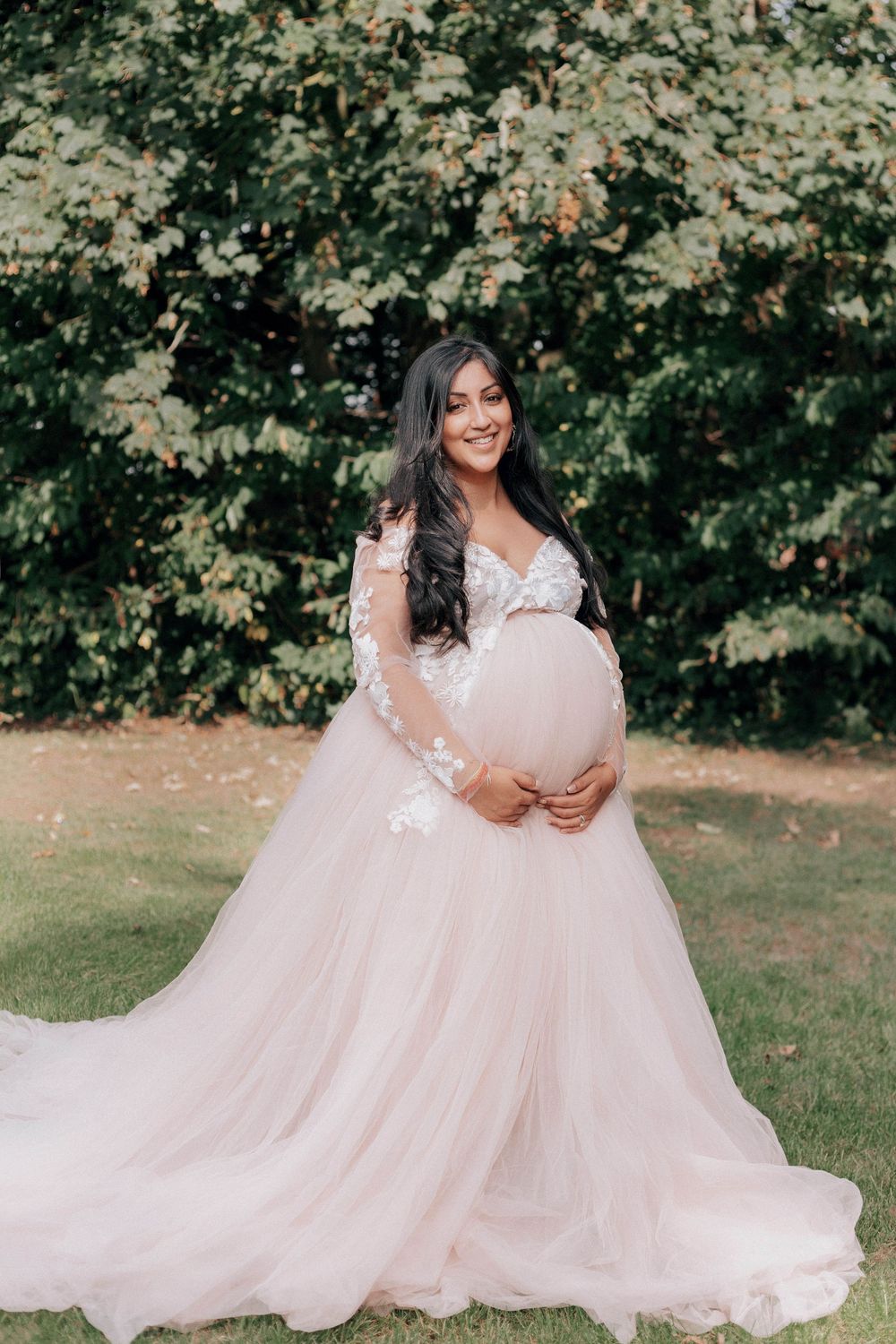 Pregnant woman in blush pink lace tulle maternity gown cradling baby bump outdoors in lush green garden.