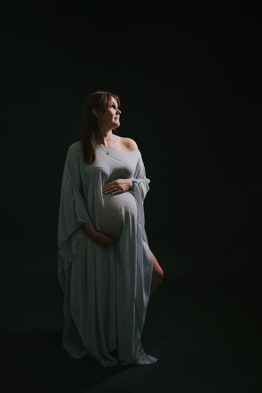 Pregnant woman in flowing white gown cradling baby bump against dark studio background.