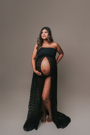 Pregnant woman in elegant black lace maternity gown posing gracefully in a professional studio photo shoot.