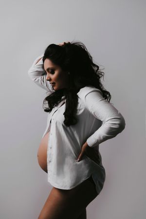 Pregnant woman in white open shirt posing sideways, showing baby bump, with hand on hip against gray background.