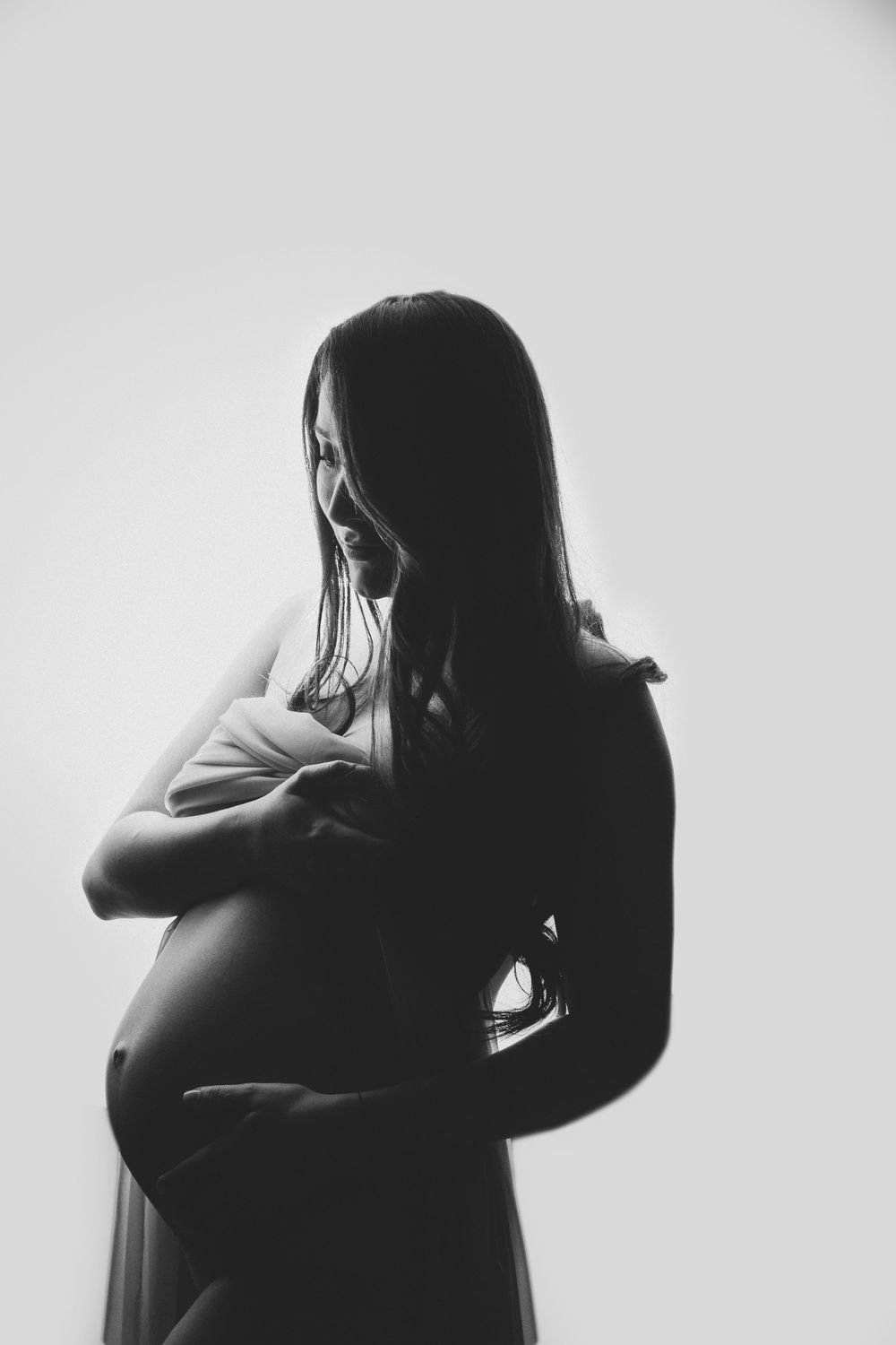 Black and white maternity photo of pregnant woman cradling her baby bump, smiling softly against light background.