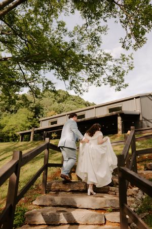 Bride and groom walking up stone steps toward a rustic venue surrounded by lush green trees on their wedding day.