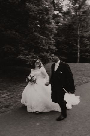 Black and white photo of a smiling bride in lace gown and groom in tuxedo walking hand in hand on a tree-lined path.