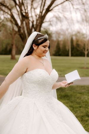 Bride in white ballgown and tiara reads a note outdoors, smiling softly amid lush green trees.