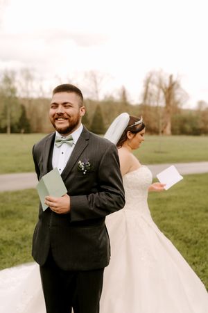 Smiling groom in gray suit reads letter back-to-back with bride in white gown and veil on green lawn.