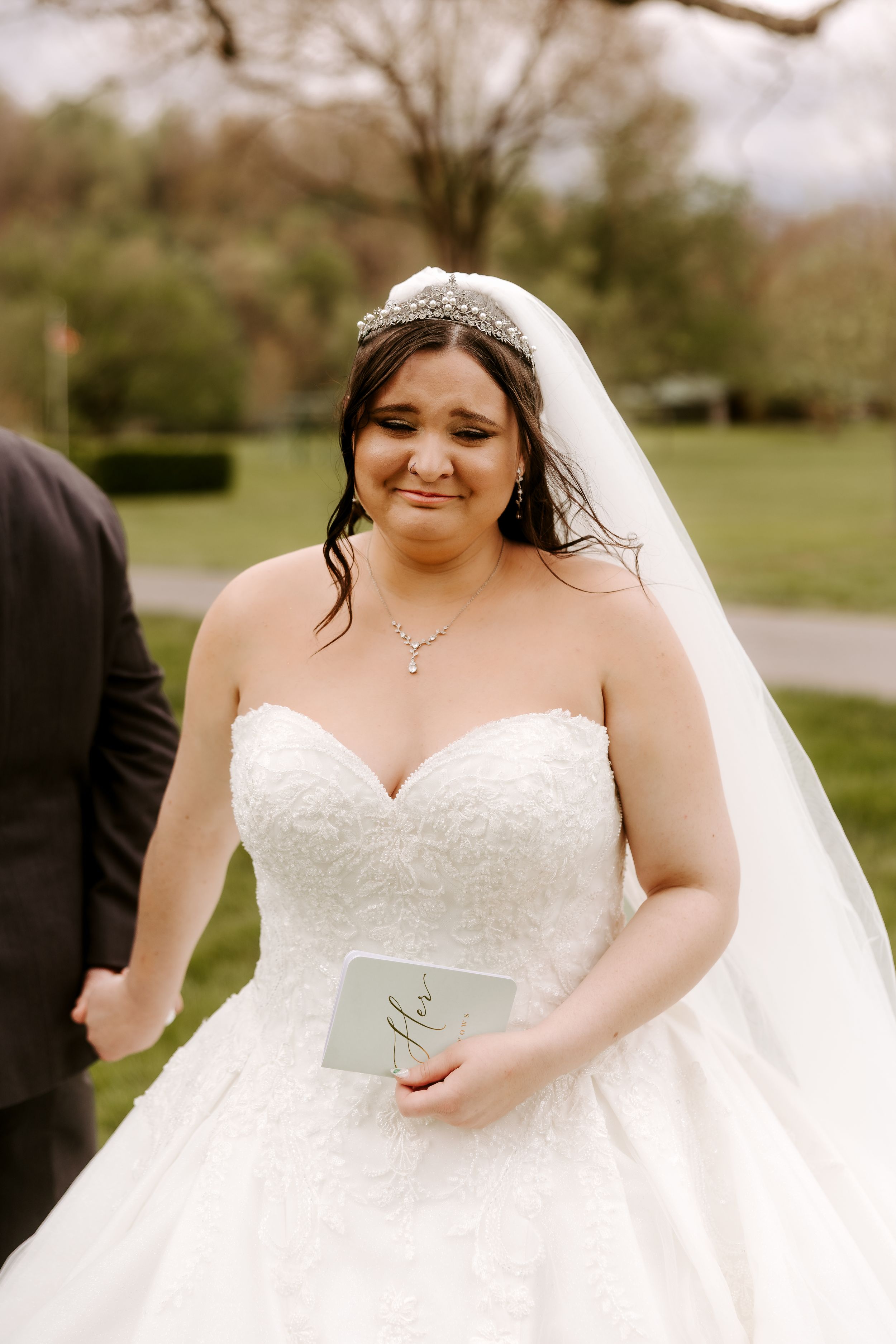 Bride in strapless lace ballgown with veil and pearl headpiece smiles while holding vows card outdoors.