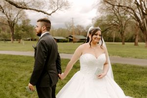 Bride in white ballgown holds hands with groom in black suit walking through green park on wedding day.