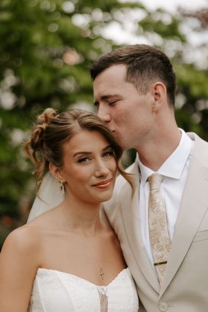 Groom in beige suit kissing bride on temple, bride smiling in white strapless gown with veil outdoors.