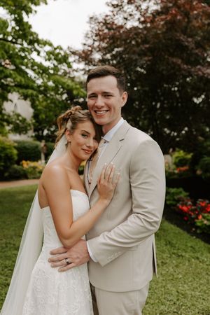 A smiling bride and groom pose together outdoors in a lush garden, she in a white gown and he in a beige suit.