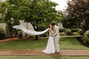 Bride and groom kissing outdoors with veil flowing in wind, lush green trees and white building backdrop.