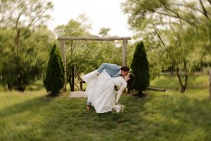 Bride and groom share a romantic dip kiss on lush green lawn framed by wooden arch and evergreen trees.
