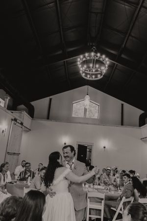 Black and white photo of a couple sharing their first dance at a rustic barn wedding reception with chandelier lighting.