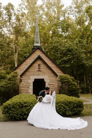 Bride and groom kissing in front of a rustic stone chapel surrounded by lush green trees and bushes.