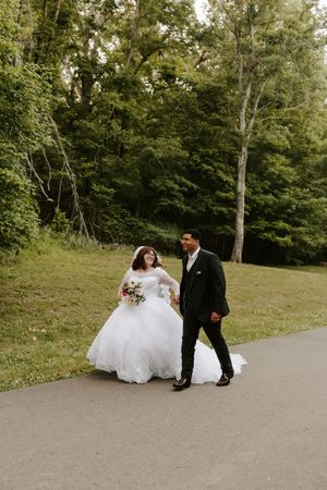 Bride in white ball gown and groom in dark suit walk hand-in-hand on a path surrounded by lush green trees.