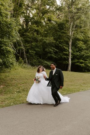 A bride in a white gown and groom in a dark suit walk hand-in-hand on a path surrounded by lush green trees.
