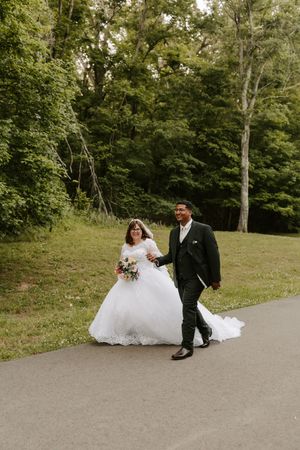 Bride in white ball gown and groom in black tuxedo walk hand in hand on a path surrounded by lush green trees.