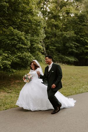 Happy bride and groom strolling along a tree-lined path, bride holding a bouquet in her white gown.