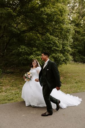 Laughing bride in white gown and groom in dark suit walk joyfully along a tree-lined path.