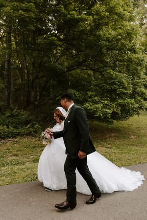 A bride and groom walk hand in hand on a path surrounded by lush green trees on their wedding day.