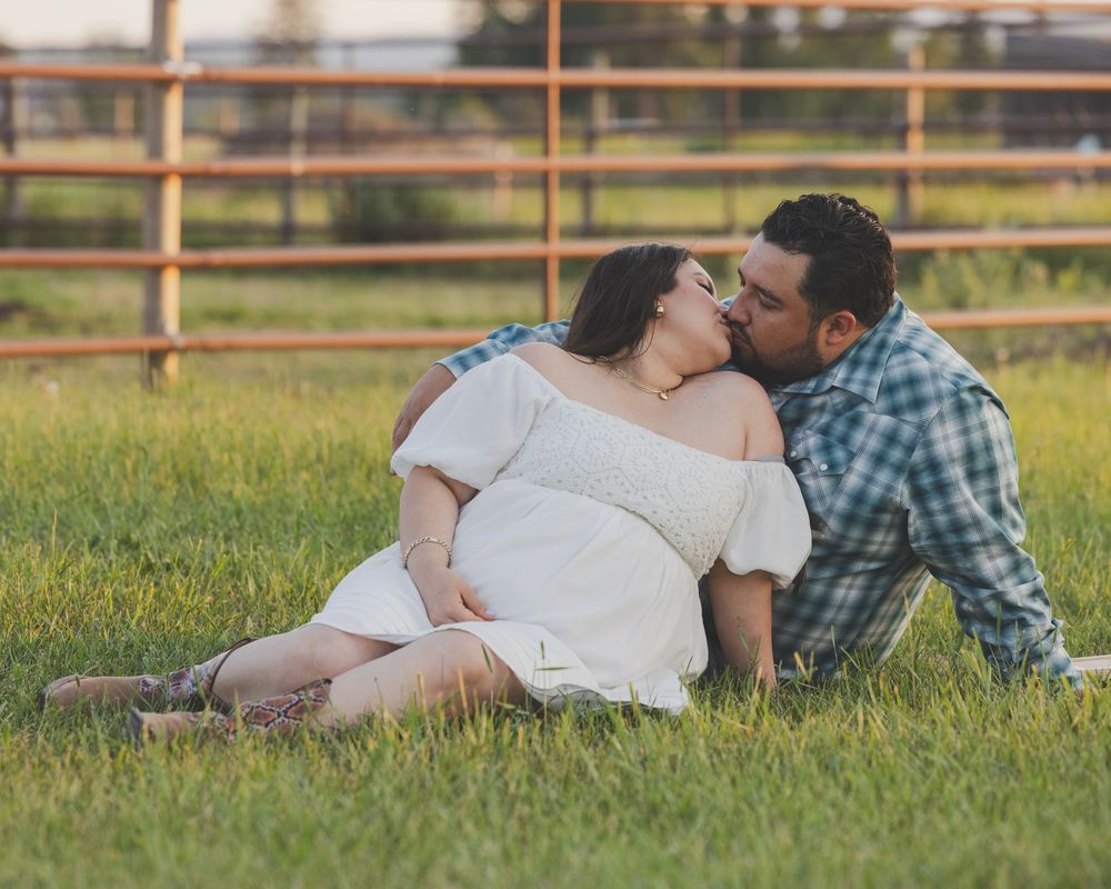 Western Couple sharing a romantic kiss while sitting in green grass field near a wooden fence during golden hour.