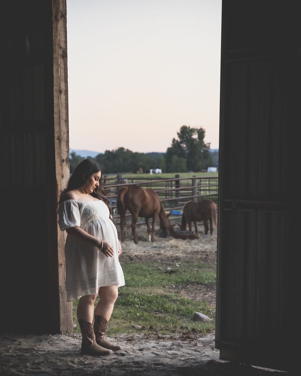 Pregnant woman in white dress and cowboy boots stands in barn doorway with horses grazing in background at dusk.
