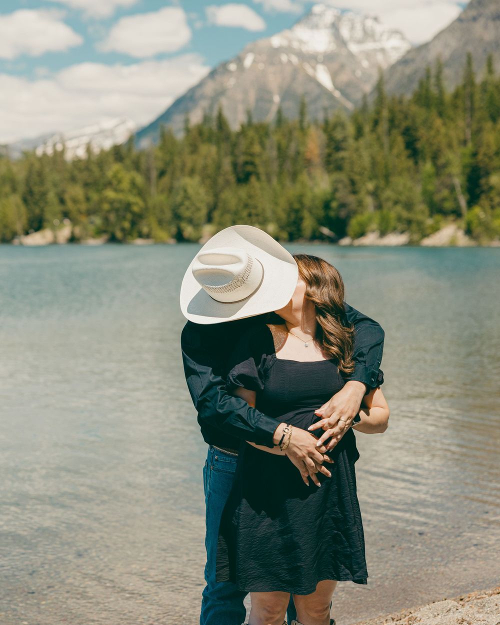 Couple sharing a romantic kiss by a mountain lake, woman in black dress, man in white cowboy hat, glacier national park  backdrop.