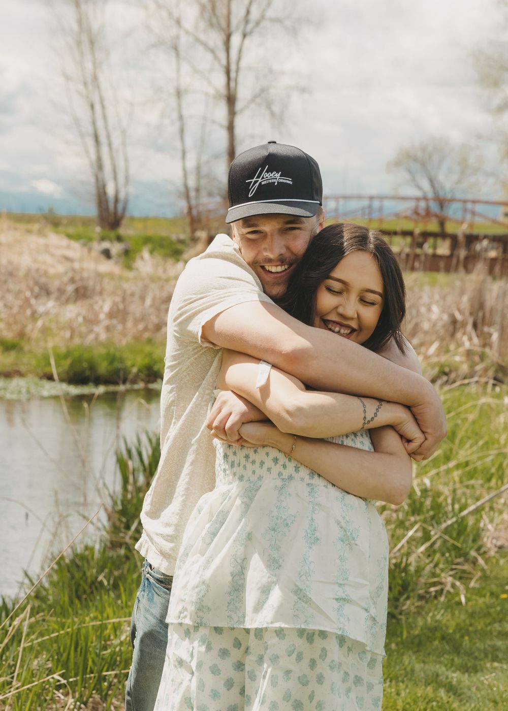 A happy couple embracing outdoors near a pond, man in snapback hat hugging smiling woman in floral dress.