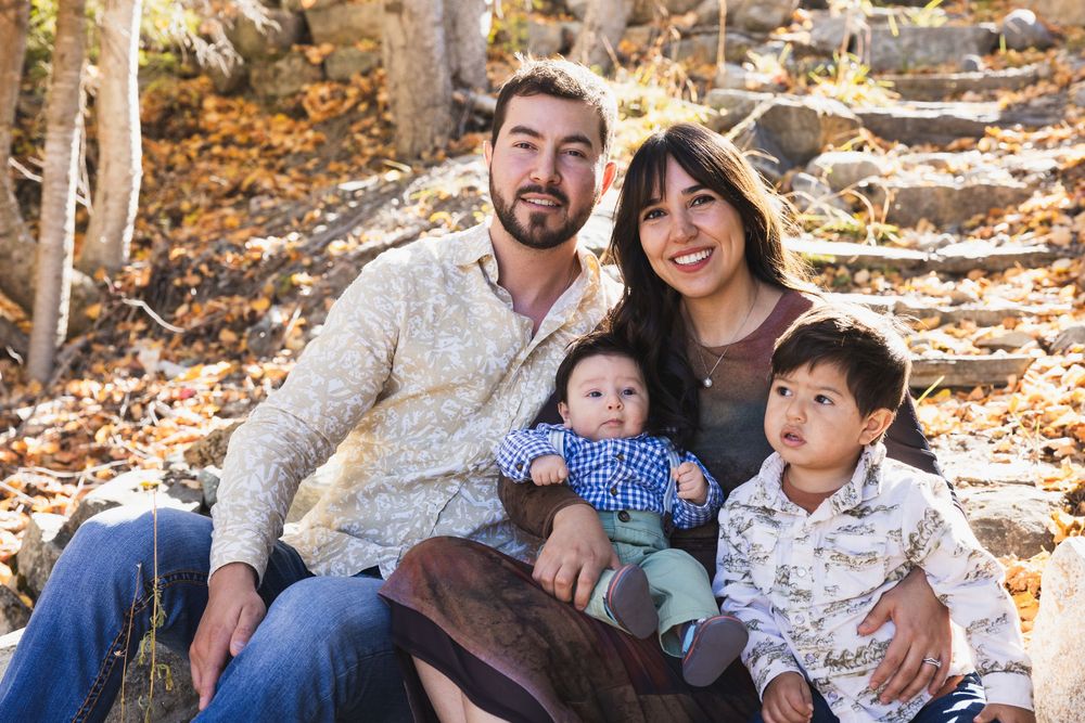Happy family of four posing outdoors in autumn, parents smiling with two young children among fall foliage.