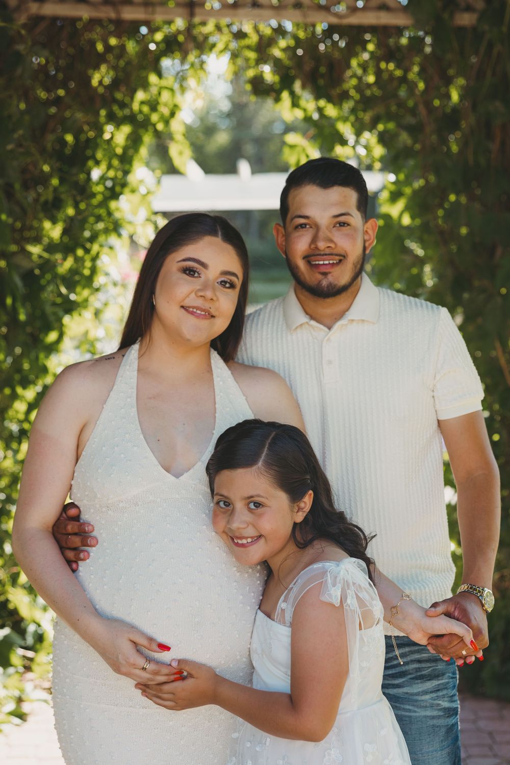 Pregnant woman in white dress poses with husband and daughter in lush green garden setting.
