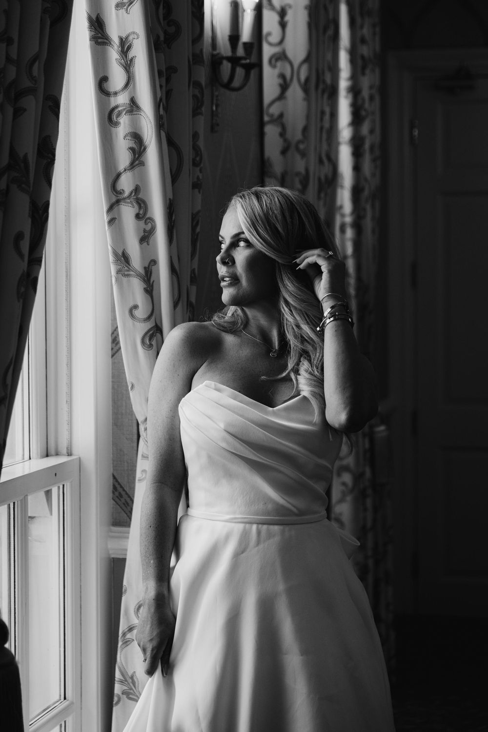 Black and white photo of a bride in a strapless gown gazing out a window with elegant curtains.