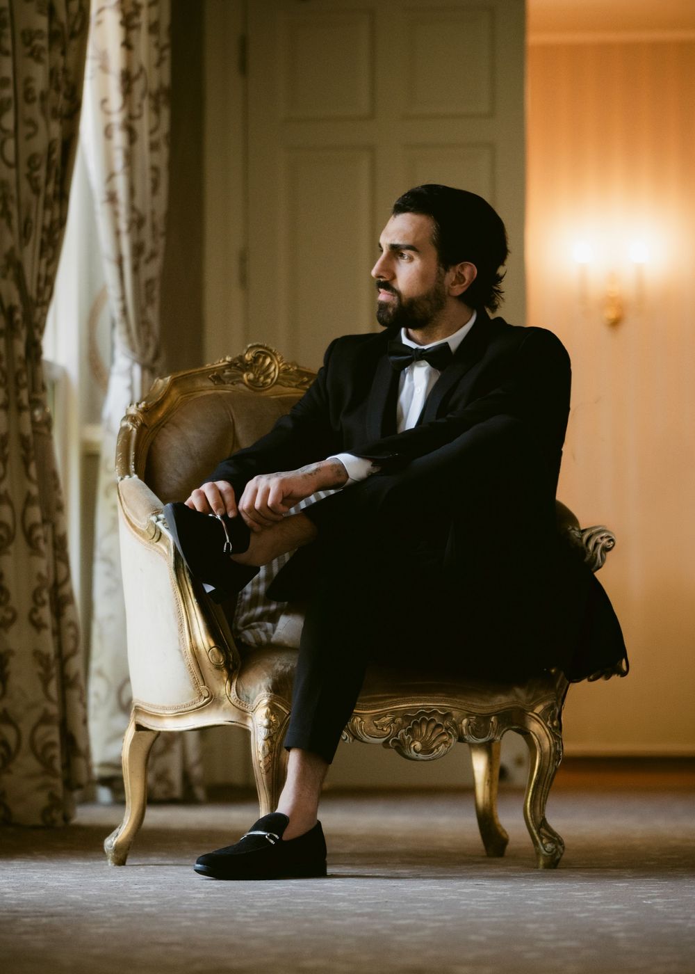 Elegant groom in black tuxedo sitting in ornate gold chair, gazing thoughtfully in luxurious room.