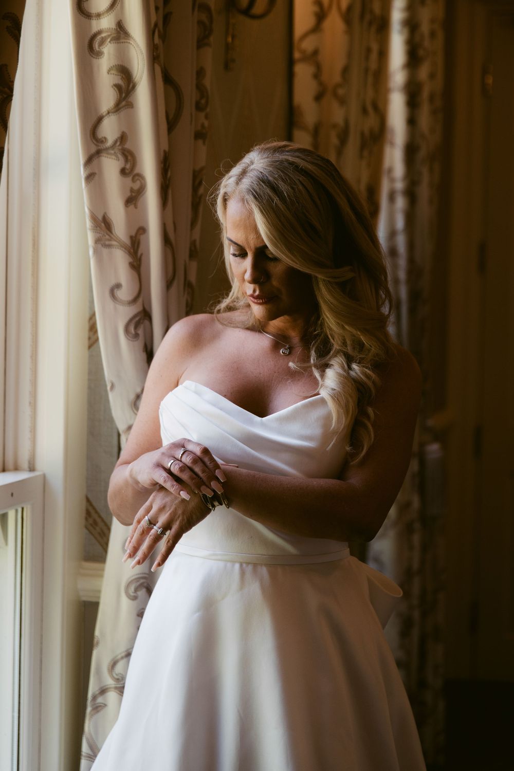 Bride in strapless white wedding gown stands by window, arms crossed, looking down in elegant bridal portrait.