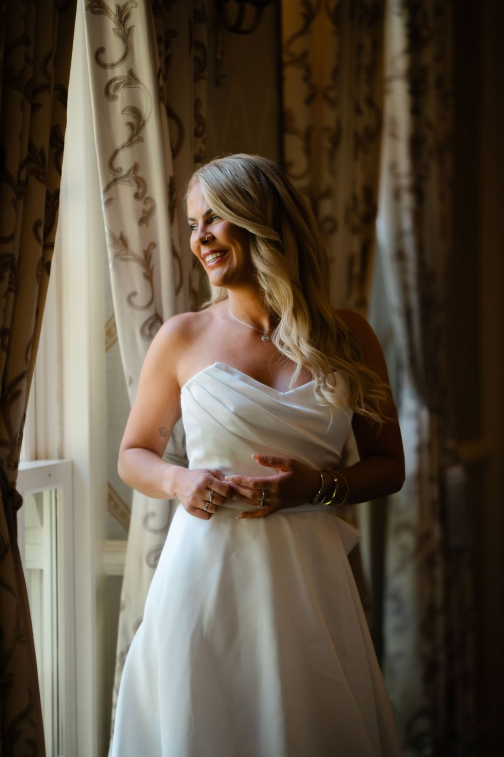 Smiling bride in strapless white wedding gown standing by draped curtained window in soft natural light.