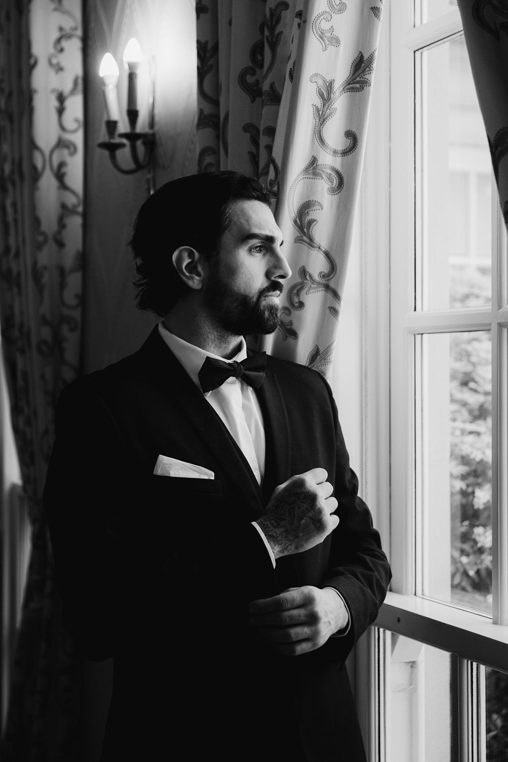 Black and white portrait of a bearded man in a tuxedo with bow tie gazing out a window in an elegant room.