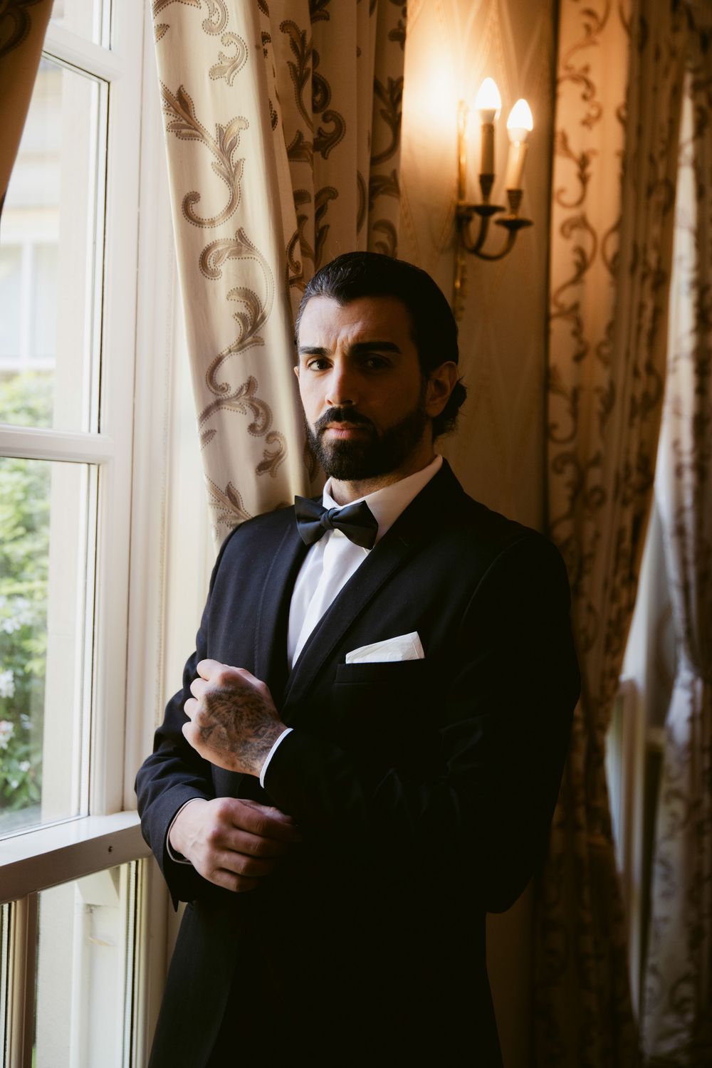 Bearded groom in black tuxedo and bow tie adjusting cufflinks by a window with elegant curtains.
