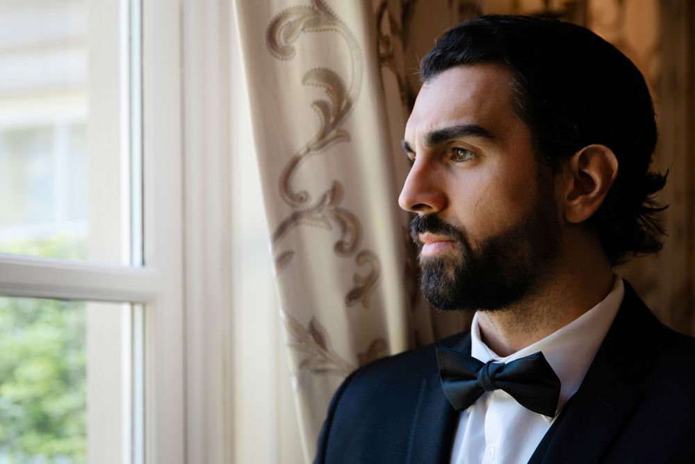 Bearded groom in navy suit and bow tie gazing thoughtfully out a window on his wedding day.