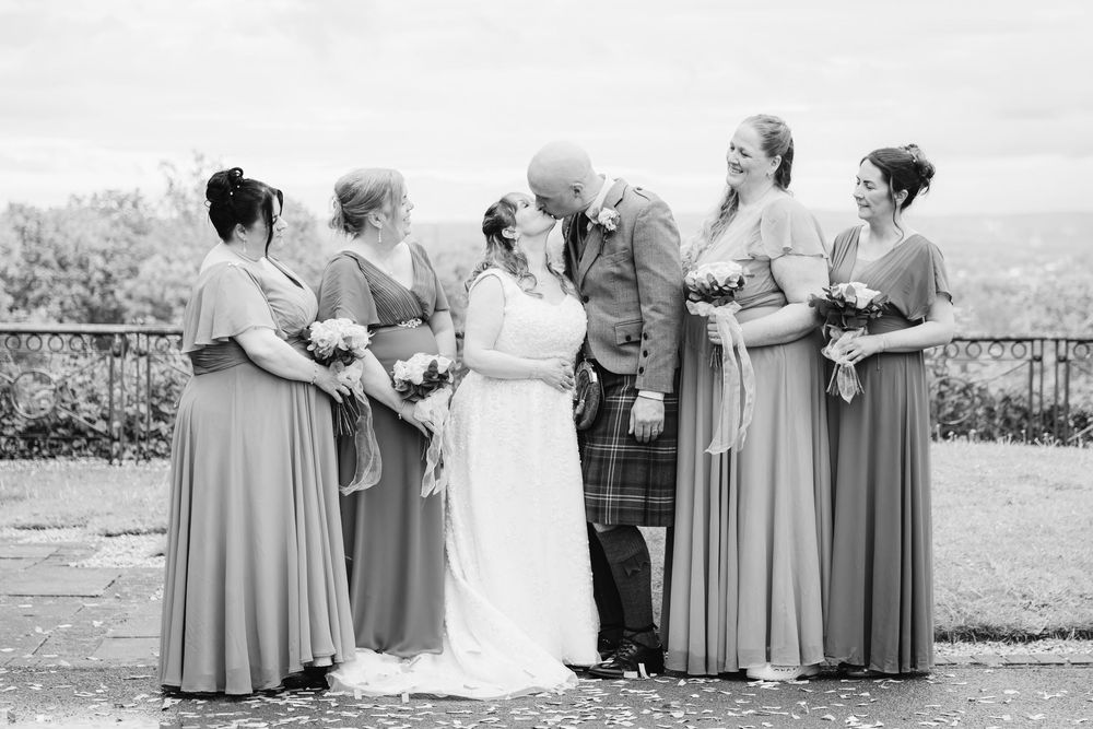 Bride and groom kissing surrounded by bridesmaids in long dresses at an outdoor wedding ceremony in Scotland.