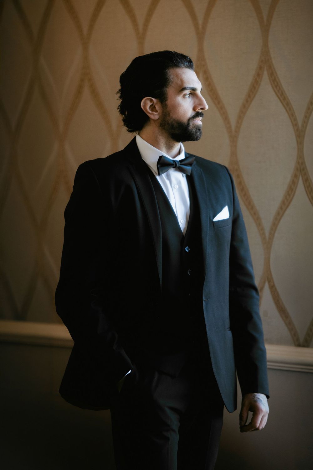 Groom in black tuxedo with bow tie and white pocket square standing elegantly against patterned wall.