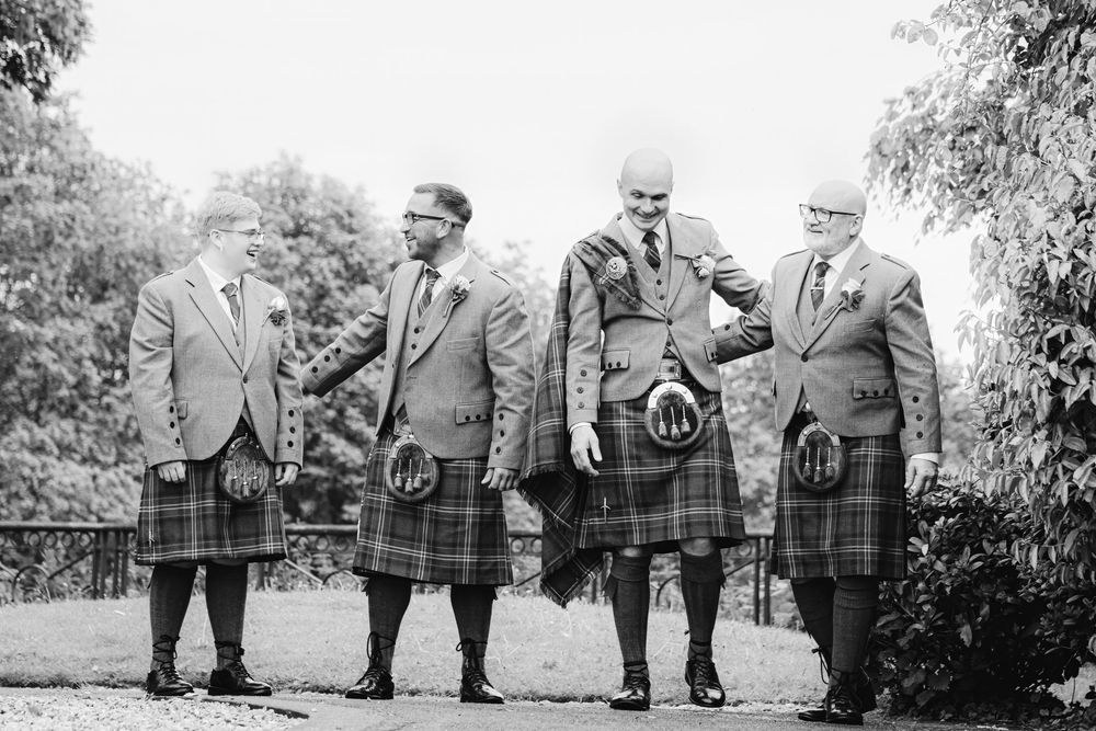 Four men in traditional Scottish kilts and jackets pose outdoors in a black and white wedding photo.