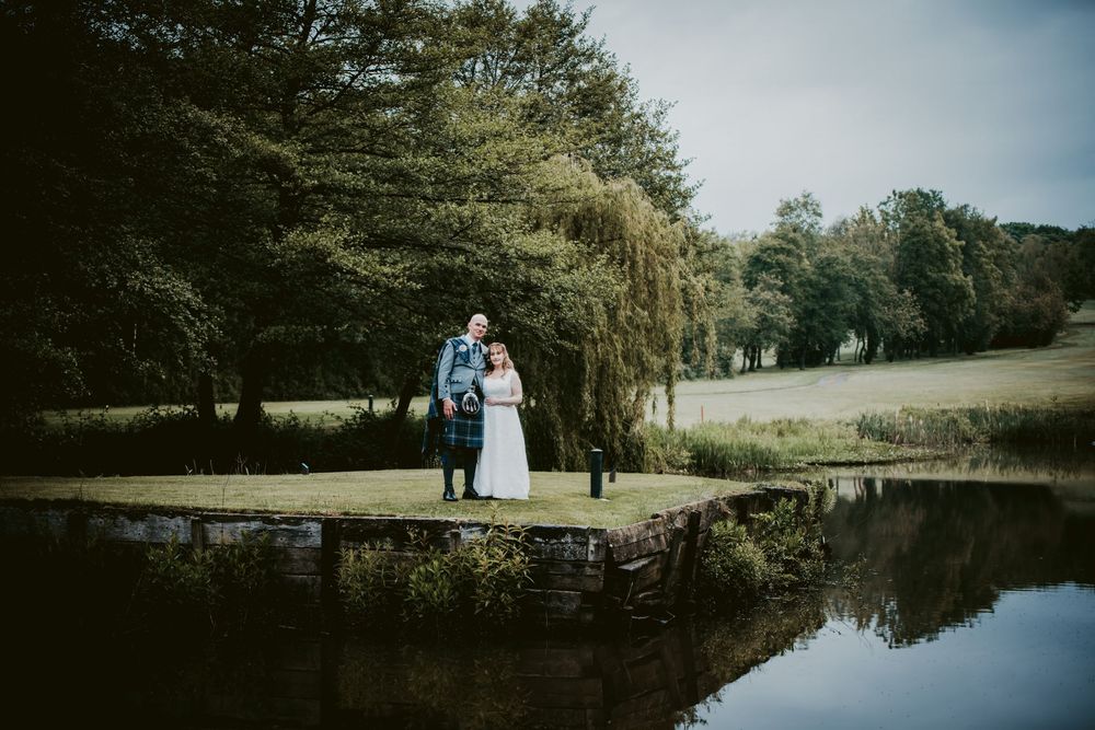 Bride and groom stand romantically by a serene pond surrounded by lush trees in a picturesque outdoor wedding setting.