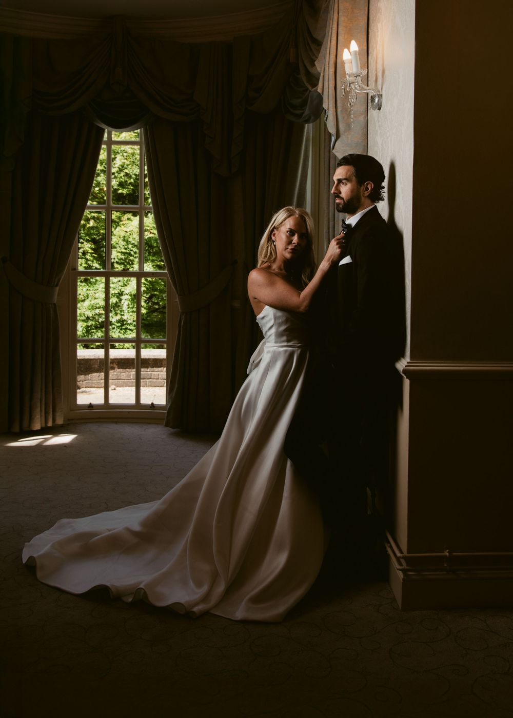 Bride and groom pose elegantly in a grand room with draped curtains and natural window light.