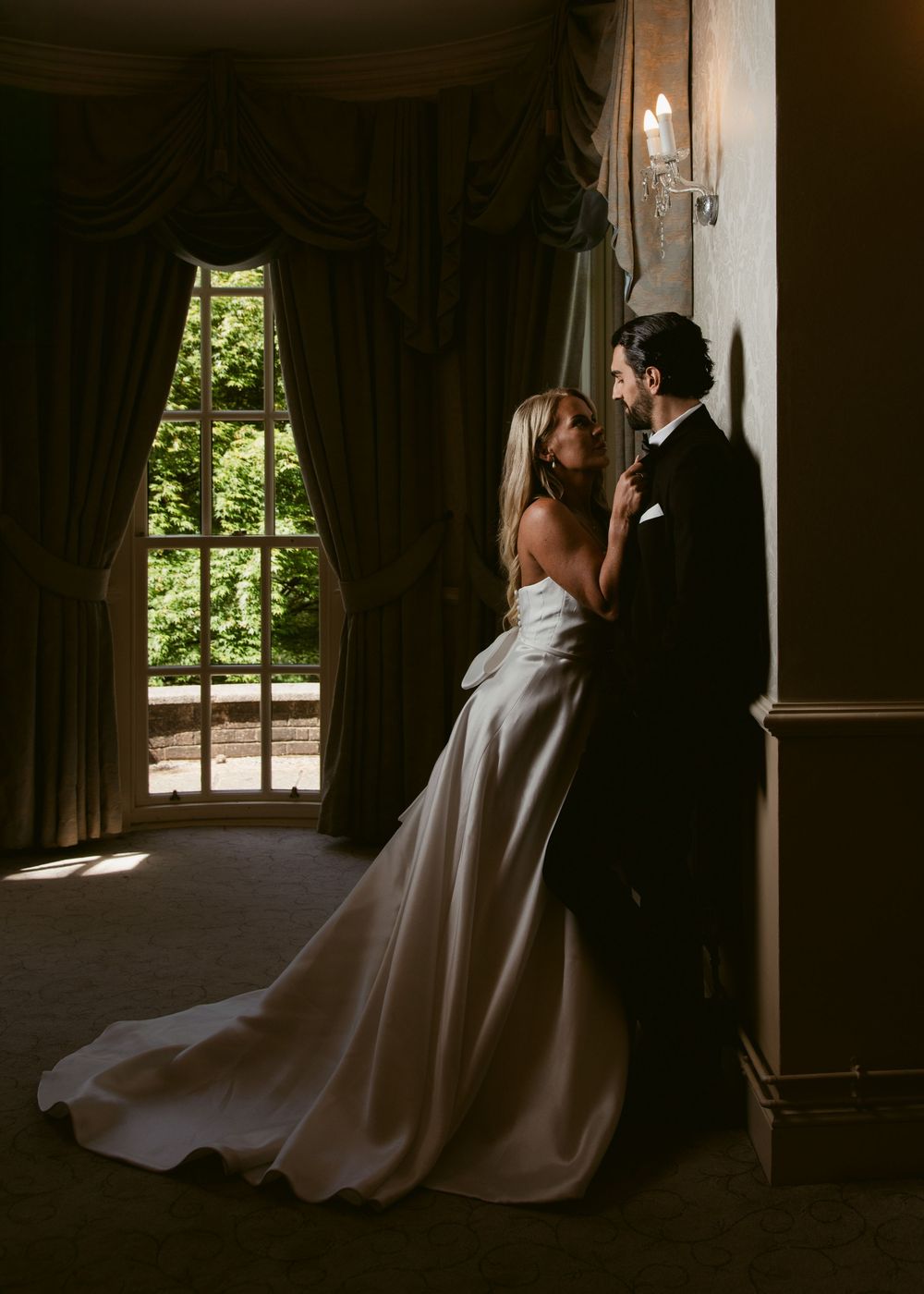 Bride and groom share a romantic moment by a sunlit window in a grand, dimly lit room.
