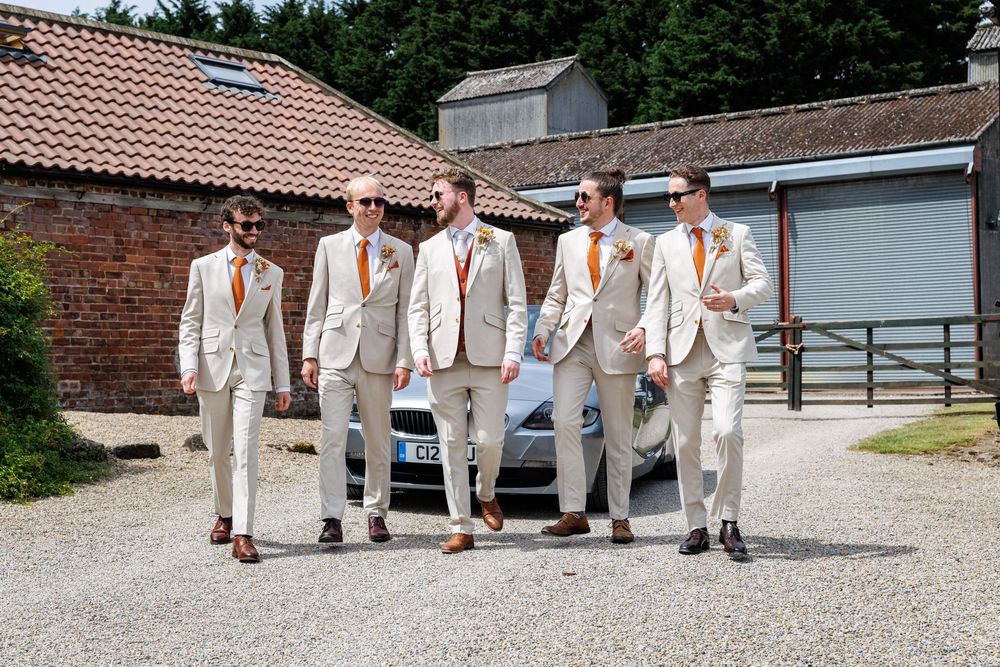 Five groomsmen in matching cream suits with orange ties walk confidently together outdoors at a rustic wedding venue.
