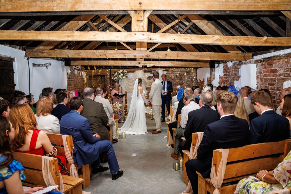 Rustic barn wedding ceremony with guests seated on wooden benches as couple exchanges vows beneath exposed timber beams.