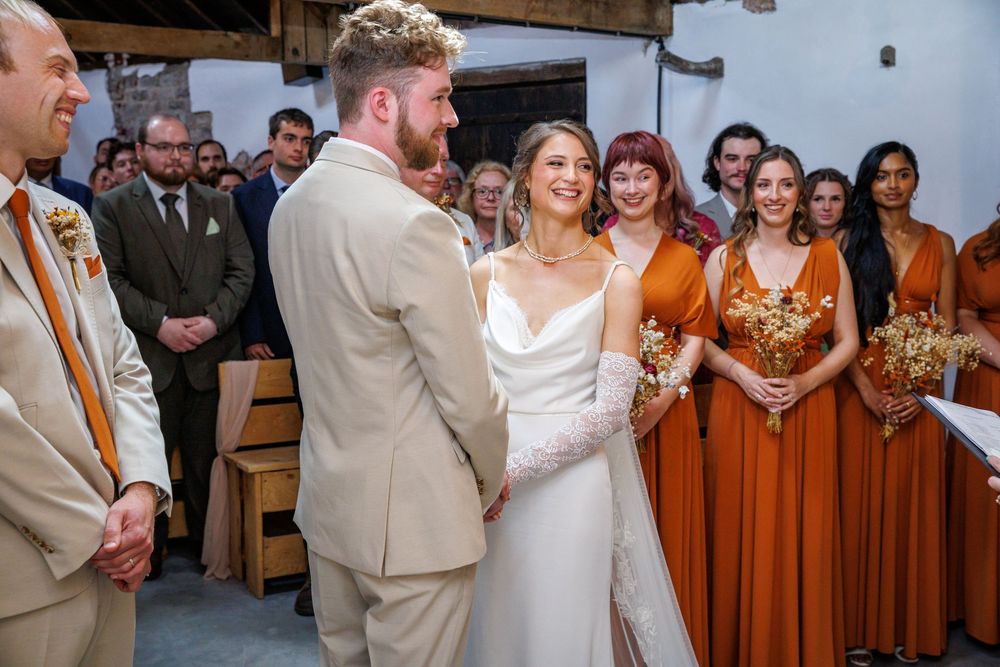 Bride and groom exchange vows at wedding ceremony with bridesmaids in rust orange dresses and guests seated in background.