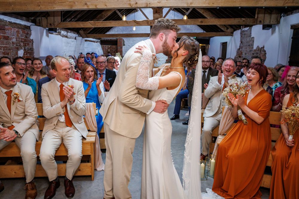 Bride and groom share first kiss at rustic barn wedding ceremony while guests applaud in background.